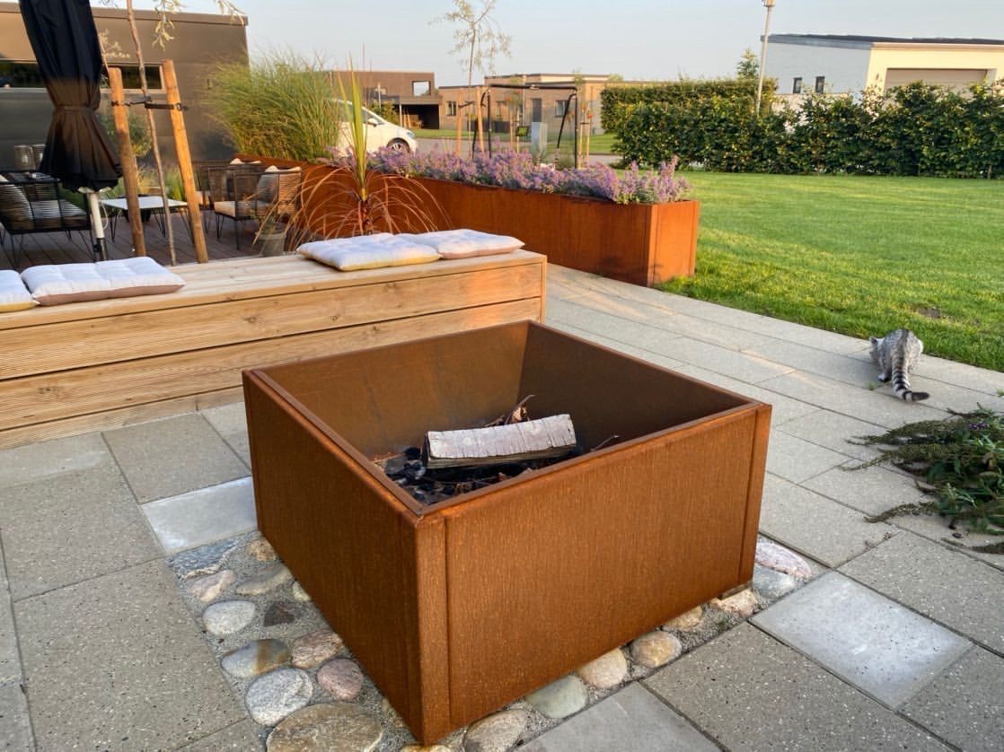 Fyrkantig eldstad i cortenstål placerad i trädgård, omgiven av uteplats och planteringar