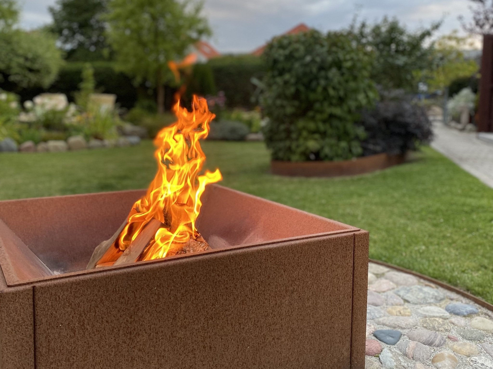 Fyrkantig eldstad i cortenstål med levande lågor placerad i grön trädgårdsmiljö