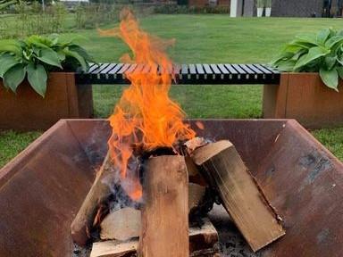 Fyrkantig eldstad i cortenstål med vedträn och öppen eld placerad i trädgård