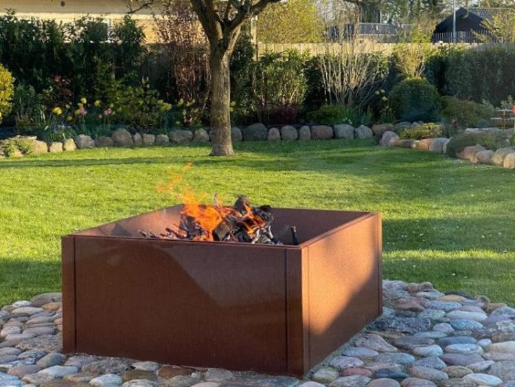 Fyrkantig eldstad i cortenstål med öppen eld placerad på stenlagd yta i trädgård
