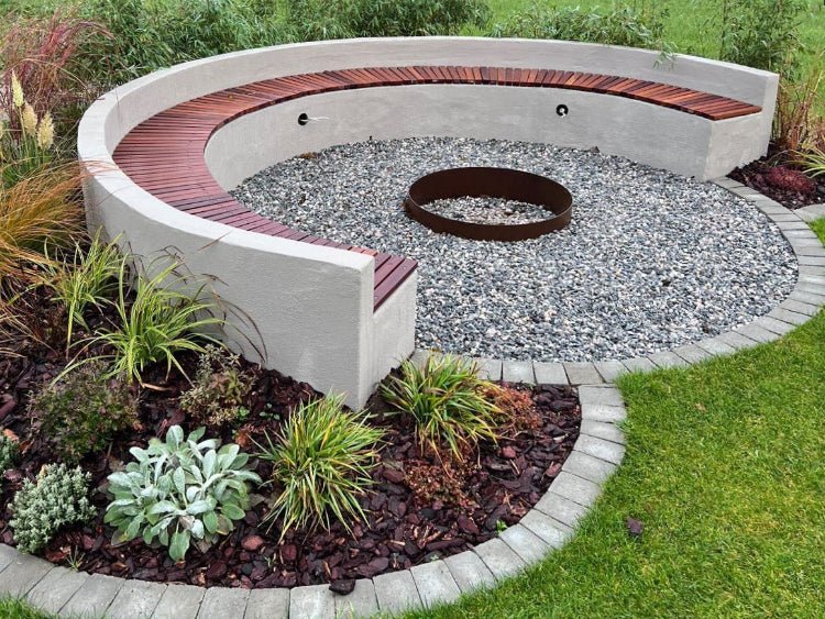 Rund eldstad i cortenstål placerad i cirkulär sittplats med bänk, grus och planteringar i trädgård