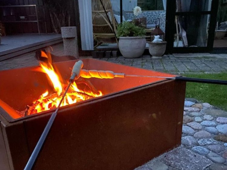 Eldstad i cortenstål med öppen eld där bröd grillas över glöden i trädgårdsmiljö