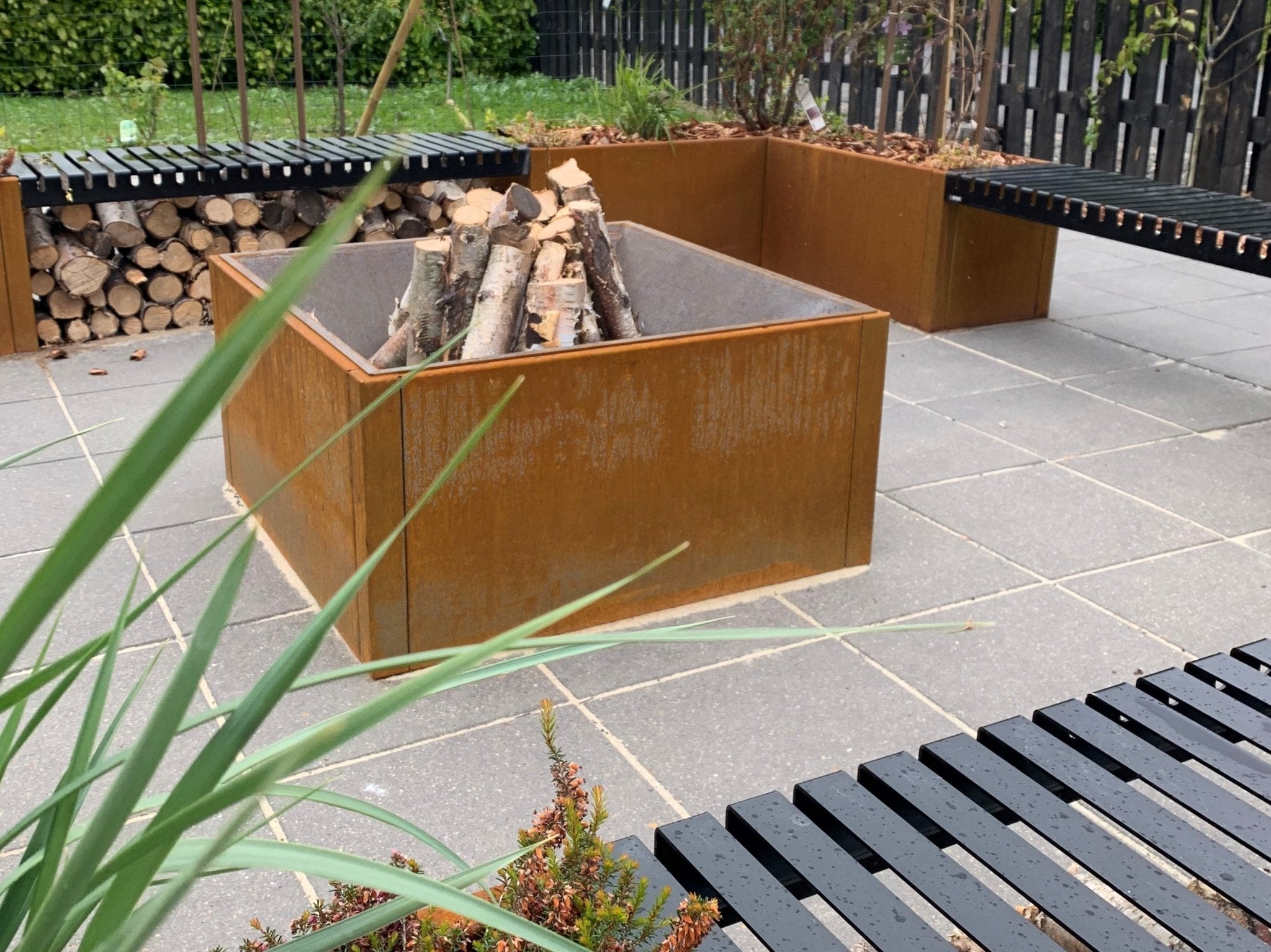 Fyrkantig eldstad i cortenstål med ved, kombinerad med integrerade sittbänkar och modern trädgårdsmiljö