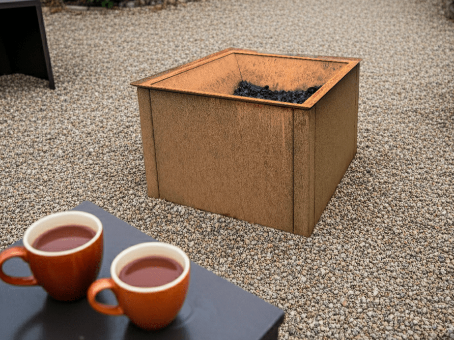 Fyrkantig eldstad i cortenstål placerad på grus, med glödbädd och kaffekoppar i förgrunden