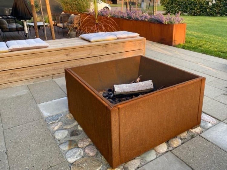 Fyrkantig eldstad i cortenstål placerad på stenläggning vid uteplats med bänk och planteringar