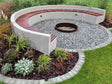 Rund eldstad i cortenstål placerad i cirkulär sittplats med bänk, grus och planteringar i trädgård